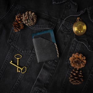 Puede incluir: Una cartera de cuero negro con detalles azules, un reloj de bolsillo dorado, dos llaves doradas y pi&ntilde;as descansan sobre una chaqueta vaquera negra. La cartera tiene un borde cosido.