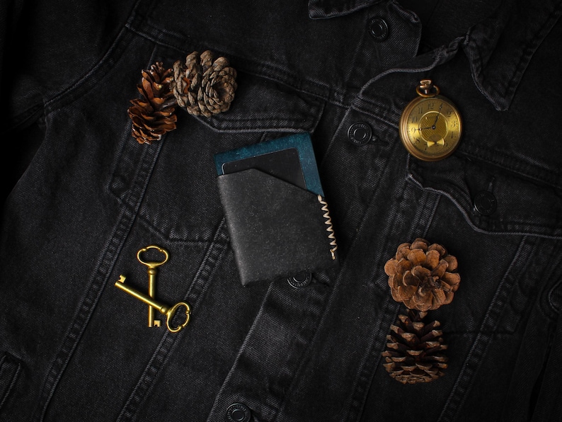 Puede incluir: Una cartera de cuero negro con detalles azules, un reloj de bolsillo dorado, dos llaves doradas y pi&ntilde;as descansan sobre una chaqueta vaquera negra. La cartera tiene un borde cosido.