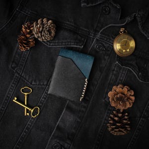 Puede incluir: Chaqueta vaquera negra con una cartera azul y negra en el bolsillo, un reloj de bolsillo dorado, dos llaves doradas y dos pi&ntilde;as. La cartera tiene un acento azul y un borde cosido.