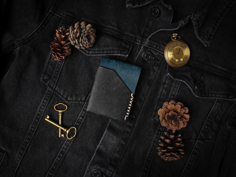 Puede incluir: Chaqueta vaquera negra con una cartera azul y negra en el bolsillo, un reloj de bolsillo dorado, dos llaves doradas y dos pi&ntilde;as. La cartera tiene un acento azul y un borde cosido.