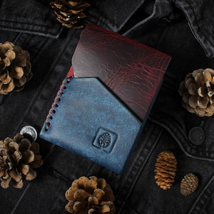 Puede incluir: Una cartera de cuero plegable en tonos azul y burdeos, con costuras rojas en el borde. La cartera presenta un logotipo de &aacute;rbol y est&aacute; rodeada de pi&ntilde;as sobre una chaqueta vaquera negra.