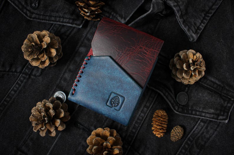 Puede incluir: Una cartera de cuero plegable en tonos azul y burdeos, con costuras rojas en el borde. La cartera presenta un logotipo de &aacute;rbol y est&aacute; rodeada de pi&ntilde;as sobre una chaqueta vaquera negra.