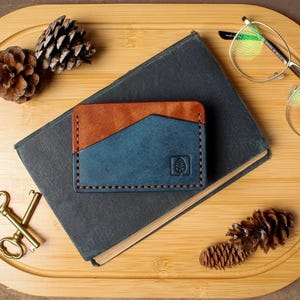May include: A handcrafted leather card wallet in brown and blue, with a tree logo, rests on a dark book. The wallet is next to a pair of eyeglasses, pine cones, and antique keys on a wooden tray.