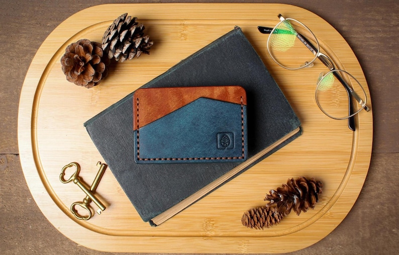May include: A handcrafted leather card wallet in brown and blue, with a tree logo, rests on a dark book. The wallet is next to a pair of eyeglasses, pine cones, and antique keys on a wooden tray.