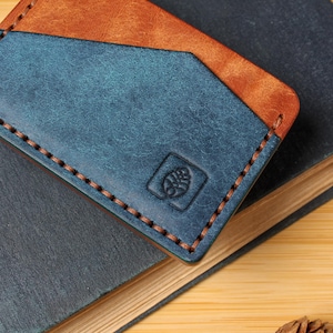 May include: A handmade leather cardholder in blue and brown, with a stitched border. The cardholder is embossed with a small emblem and rests on a dark blue book. The background is a light wood surface.