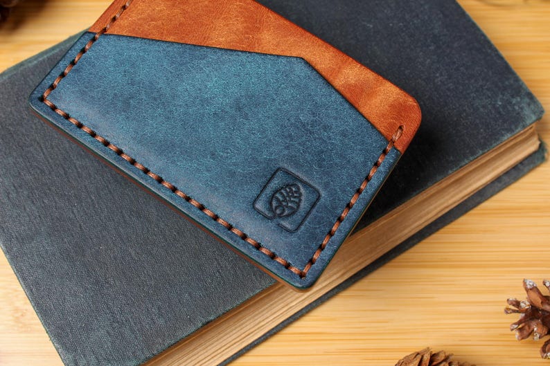 May include: A handmade leather cardholder in blue and brown, with a stitched border. The cardholder is embossed with a small emblem and rests on a dark blue book. The background is a light wood surface.