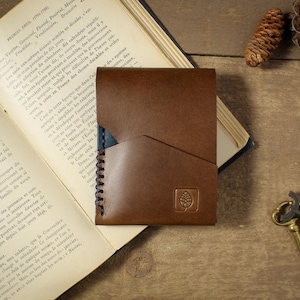 May include: A brown leather card holder featuring a diagonal pocket design, dark stitching along the side, and a small square logo embossed on the front. The wallet is constructed with smooth leather and displays a clean, functional aesthetic.