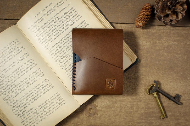 May include: A brown leather card holder featuring a diagonal pocket design, dark stitching along the side, and a small square logo embossed on the front. The wallet is constructed with smooth leather and displays a clean, functional aesthetic.