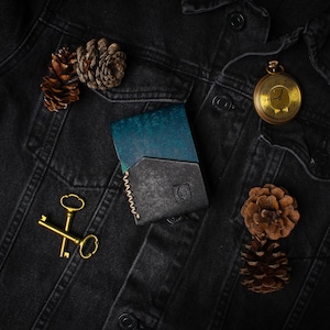 Puede incluir: Cartera de cuero bicolor, turquesa y negro, sobre una chaqueta vaquera negra. Un reloj de bolsillo dorado, dos llaves doradas y pi&ntilde;as completan la composici&oacute;n. La cartera tiene un borde cosido.