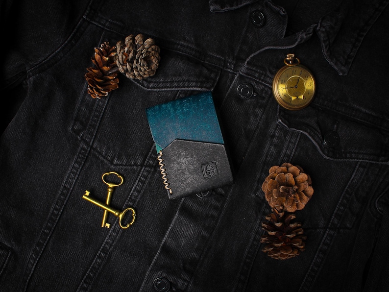Puede incluir: Cartera de cuero bicolor, turquesa y negro, sobre una chaqueta vaquera negra. Un reloj de bolsillo dorado, dos llaves doradas y pi&ntilde;as completan la composici&oacute;n. La cartera tiene un borde cosido.