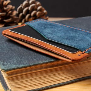 May include: A handmade leather cardholder in shades of blue and brown, with visible stitching. The cardholder is placed on a vintage book with aged pages. The design includes a blue suede panel and a contrasting brown leather base.