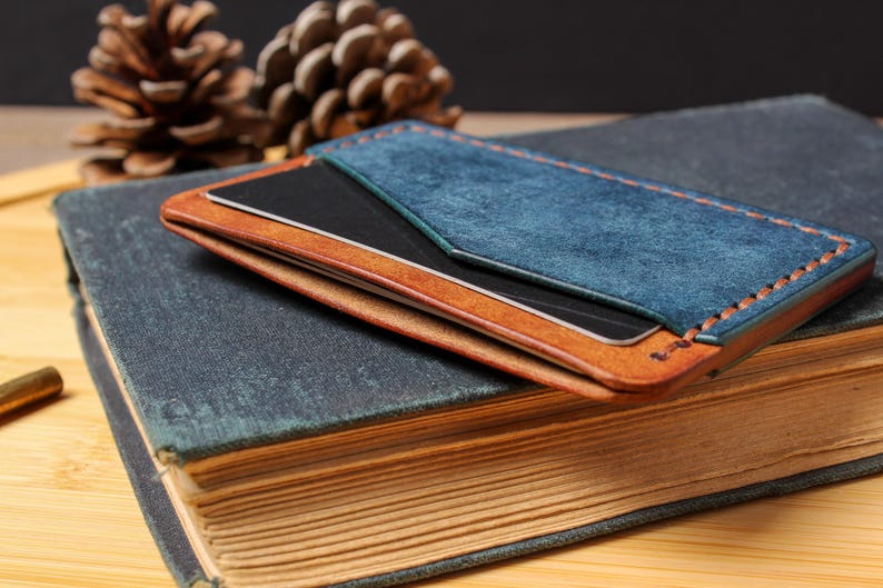 May include: A handmade leather cardholder in shades of blue and brown, with visible stitching. The cardholder is placed on a vintage book with aged pages. The design includes a blue suede panel and a contrasting brown leather base.