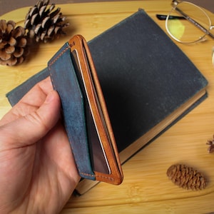 May include: A blue and brown leather wallet with a minimalist design. The wallet is held in a hand and is set against a backdrop of a book, glasses, and pine cones. The wallet has a bi-fold design.