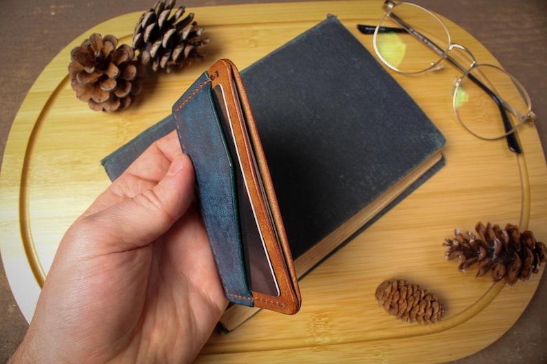 May include: A blue and brown leather wallet with a minimalist design. The wallet is held in a hand and is set against a backdrop of a book, glasses, and pine cones. The wallet has a bi-fold design.