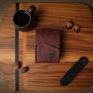 Puede incluir: Una cartera de cuero marr&oacute;n con un logotipo de &aacute;rbol, una navaja plegable negra y una peque&ntilde;a taza de bronce llena de l&iacute;quido oscuro est&aacute;n dispuestas sobre una tabla de cortar de madera. Dos bellotas y dos pi&ntilde;as tambi&eacute;n est&aacute;n en la tabla.