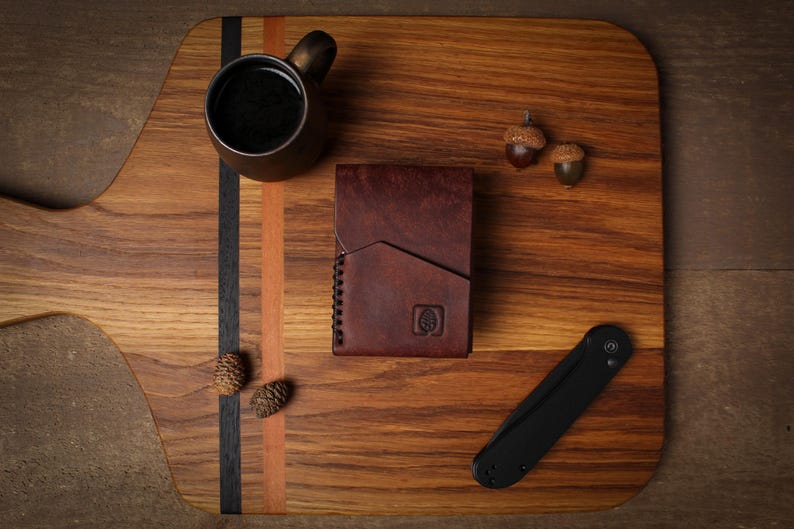 Puede incluir: Una cartera de cuero marr&oacute;n con un logotipo de &aacute;rbol, una navaja plegable negra y una peque&ntilde;a taza de bronce llena de l&iacute;quido oscuro est&aacute;n dispuestas sobre una tabla de cortar de madera. Dos bellotas y dos pi&ntilde;as tambi&eacute;n est&aacute;n en la tabla.