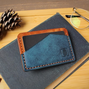 May include: A handmade leather card holder in blue and brown, with a stitched design and a pinecone emblem. The wallet is on a dark book, with pinecones and glasses in the background.