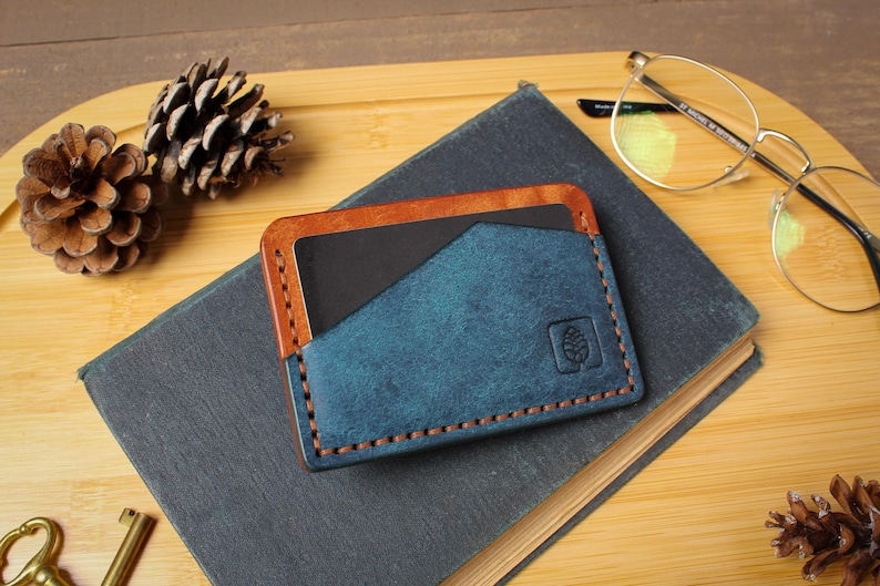 May include: A handmade leather card holder in blue and brown, with a stitched design and a pinecone emblem. The wallet is on a dark book, with pinecones and glasses in the background.