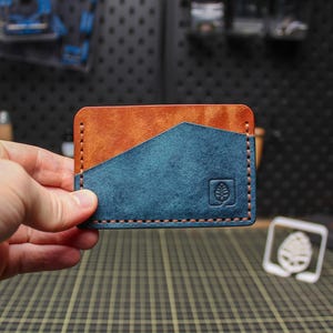 May include: A handcrafted leather card wallet with a two-tone design. The top section is a warm brown, while the lower section is a deep teal. The wallet features a stitched border and a debossed leaf logo.
