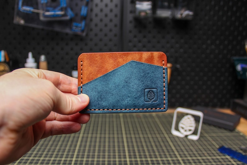 May include: A handcrafted leather card wallet with a two-tone design. The top section is a warm brown, while the lower section is a deep teal. The wallet features a stitched border and a debossed leaf logo.