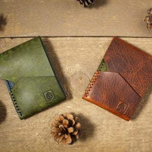 May include: Two leather wallets, one in olive green and one in reddish-brown, each featuring a diagonal front pocket, visible side stitching, and a small square logo embossed with a leaf icon on the lower right corner of the front panel.