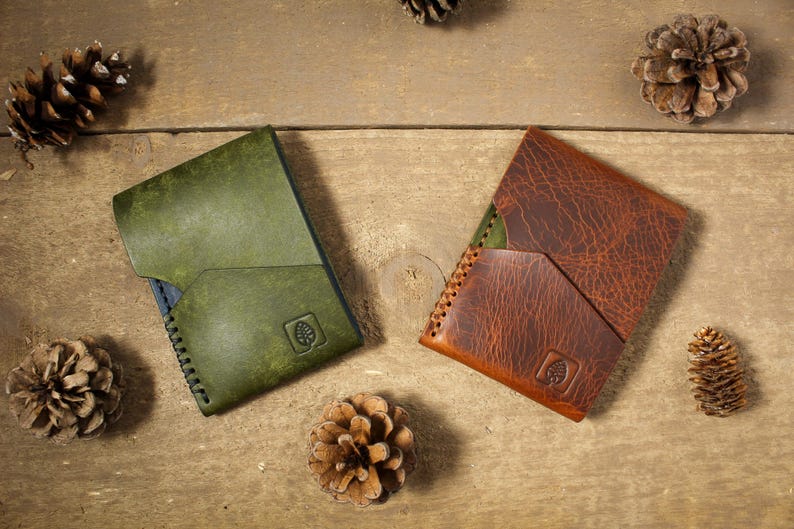 May include: Two leather wallets, one in olive green and one in reddish-brown, each featuring a diagonal front pocket, visible side stitching, and a small square logo embossed with a leaf icon on the lower right corner of the front panel.