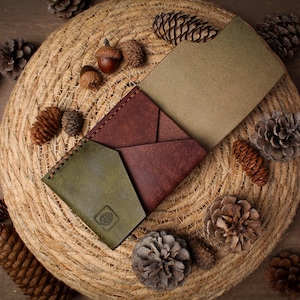 May include: A multi-colored leather wallet featuring a dark red central section with geometric folds, flanked by an olive green pocket on the left and a tan interior panel on the right. The olive green section displays a small embossed pinecone logo.