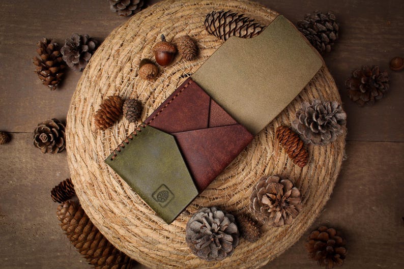 May include: A multi-colored leather wallet featuring a dark red central section with geometric folds, flanked by an olive green pocket on the left and a tan interior panel on the right. The olive green section displays a small embossed pinecone logo.