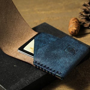 Puede incluir: Una cartera de cuero azul con una tarjeta dentro, colocada sobre una superficie oscura. La cartera tiene un acabado texturizado y est&aacute; cosida con hilo azul. Una pi&ntilde;a y una peque&ntilde;a bellota est&aacute;n en el fondo.