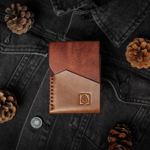 Puede incluir: Una cartera de cuero de dos tonos en marr&oacute;n y beige, con un dise&ntilde;o geom&eacute;trico y un logotipo de &aacute;rbol en relieve. La cartera est&aacute; rodeada de pi&ntilde;as en una chaqueta vaquera oscura.