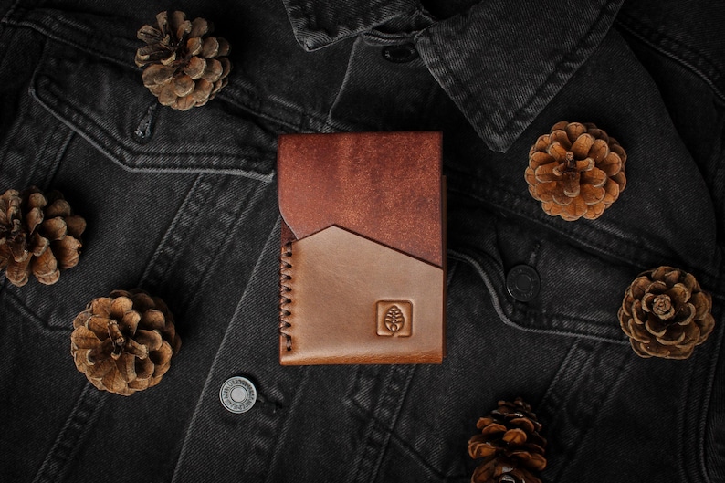 Puede incluir: Una cartera de cuero de dos tonos en marr&oacute;n y beige, con un dise&ntilde;o geom&eacute;trico y un logotipo de &aacute;rbol en relieve. La cartera est&aacute; rodeada de pi&ntilde;as en una chaqueta vaquera oscura.