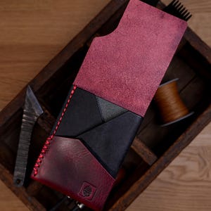 May include: A vertical leather wallet featuring a combination of deep burgundy and black panels. The wallet has a fold-over flap, red contrast stitching along the side, and a small embossed leaf logo on the bottom front corner section.