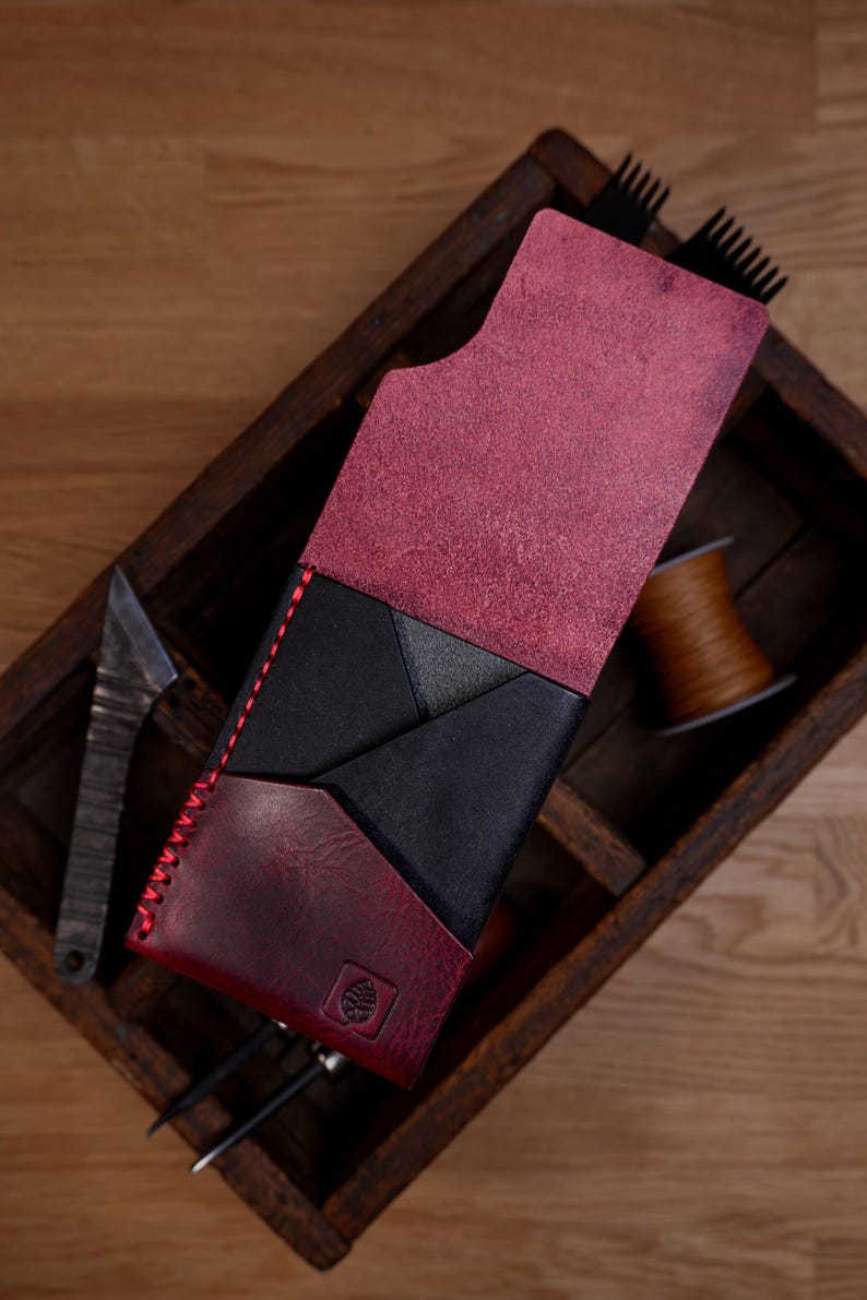May include: A vertical leather wallet featuring a combination of deep burgundy and black panels. The wallet has a fold-over flap, red contrast stitching along the side, and a small embossed leaf logo on the bottom front corner section.