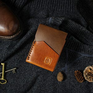 Puede incluir: Una cartera de cuero de dos tonos con un borde cosido y un emblema de pi&ntilde;a grabado. La cartera est&aacute; colocada sobre una superficie texturizada gris oscuro con un zapato de cuero marr&oacute;n, llaves y pi&ntilde;as.