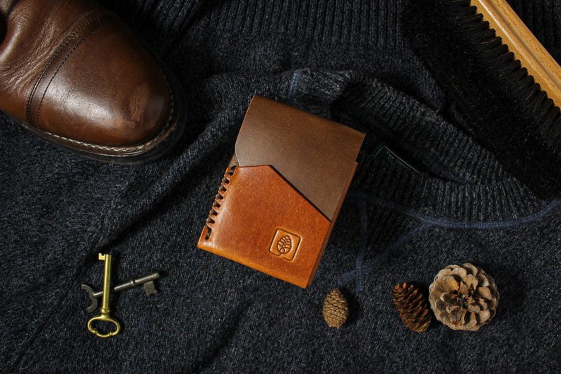 Puede incluir: Una cartera de cuero de dos tonos con un borde cosido y un emblema de pi&ntilde;a grabado. La cartera est&aacute; colocada sobre una superficie texturizada gris oscuro con un zapato de cuero marr&oacute;n, llaves y pi&ntilde;as.