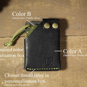 May include: A black leather wallet with a green paracord loop and a green thread stitch. The wallet has two brass rivets and the text "Color B (Shown here: Pueblo olive)" and "Color A (Shown here: Pueblo black)" is visible.