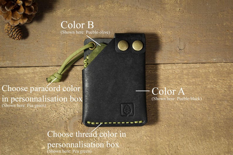 May include: A black leather wallet with a green paracord loop and a green thread stitch. The wallet has two brass rivets and the text "Color B (Shown here: Pueblo olive)" and "Color A (Shown here: Pueblo black)" is visible.