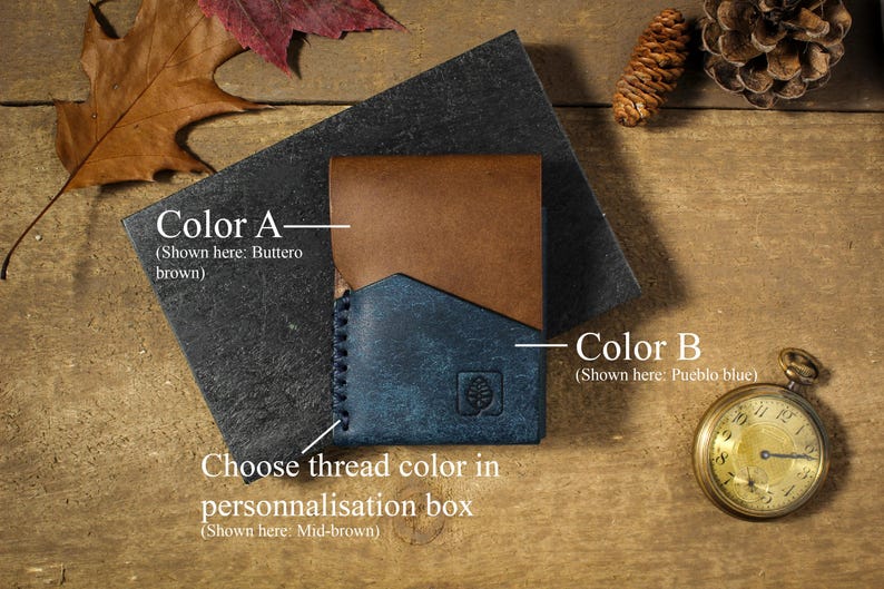 May include: A leather wallet with two different colored panels. The top panel is brown and the bottom panel is blue. The text "Color A - (Shown here: Buttero brown)" is above the top panel. The text "Color B - (Shown here: Pueblo blue)" is to the right of the bottom panel. The text "Choose thread color in personalisation box (Shown here: Mid-brown)" is below the wallet.