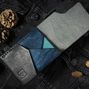 May include: A bi-fold wallet featuring a combination of textured charcoal gray and deep blue leather panels with a small triangular teal accent. The wallet has visible stitching along the edges and a small embossed square logo on the front panel.