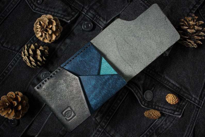 May include: A bi-fold wallet featuring a combination of textured charcoal gray and deep blue leather panels with a small triangular teal accent. The wallet has visible stitching along the edges and a small embossed square logo on the front panel.