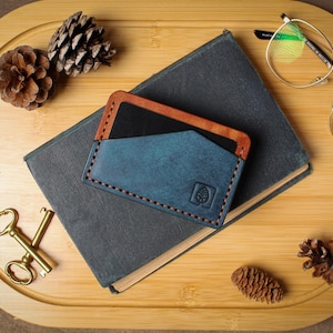 May include: A blue and brown leather card holder with a leaf logo, resting on a dark gray book. The card holder is stitched with orange thread. A pair of gold-rimmed glasses and a pair of gold scissors are also on the wooden tray.