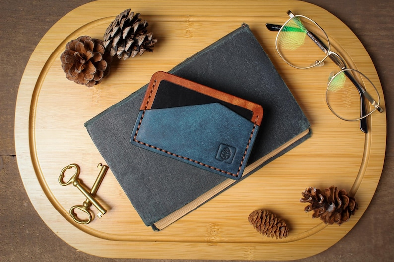 May include: A blue and brown leather card holder with a leaf logo, resting on a dark gray book. The card holder is stitched with orange thread. A pair of gold-rimmed glasses and a pair of gold scissors are also on the wooden tray.