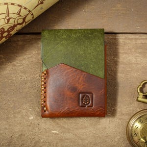 Puede incluir: Cartera de cuero hecha a mano con dise&ntilde;o bicolor. La parte superior es verde y la inferior marr&oacute;n, con un borde cosido. Se ve un peque&ntilde;o logotipo de &aacute;rbol en relieve. Un reloj de bolsillo vintage y un mapa est&aacute;n en el fondo.