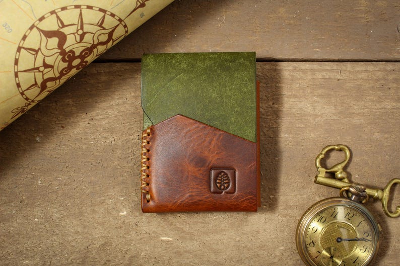 Puede incluir: Cartera de cuero hecha a mano con dise&ntilde;o bicolor. La parte superior es verde y la inferior marr&oacute;n, con un borde cosido. Se ve un peque&ntilde;o logotipo de &aacute;rbol en relieve. Un reloj de bolsillo vintage y un mapa est&aacute;n en el fondo.