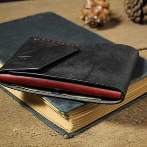 May include: A black leather wallet featuring a folded flap design with visible red stitching along the top edge. The wallet has a contrasting deep red interior layer visible along the side, and it is resting on top of a closed, dark-colored hardcover book.