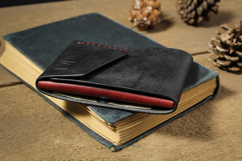 May include: A black leather wallet featuring a folded flap design with visible red stitching along the top edge. The wallet has a contrasting deep red interior layer visible along the side, and it is resting on top of a closed, dark-colored hardcover book.