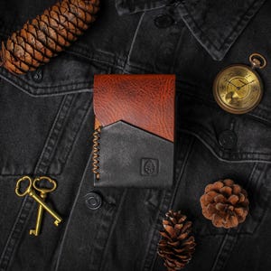 Puede incluir: Cartera de cuero bicolor marr&oacute;n y negro, con costuras en contraste y logotipo en relieve. La imagen tambi&eacute;n incluye un reloj de bolsillo vintage, llaves doradas y pi&ntilde;as, colocados sobre una chaqueta vaquera oscura.