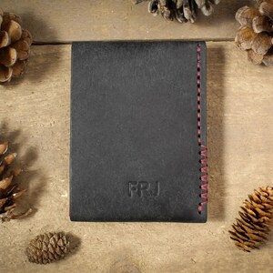 May include: A rectangular black wallet featuring the embossed letters FRJ on the front surface and a vertical row of red stitching along the right edge. The wallet is positioned centrally, surrounded by several brown pine cones.