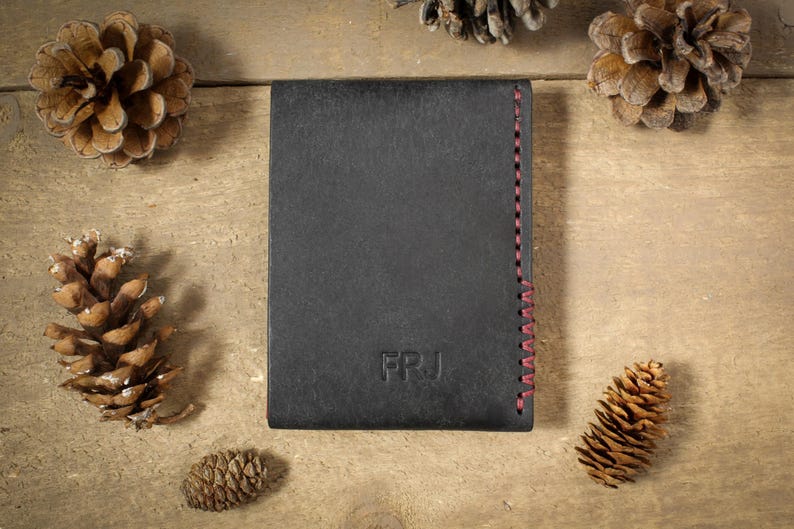 May include: A rectangular black wallet featuring the embossed letters FRJ on the front surface and a vertical row of red stitching along the right edge. The wallet is positioned centrally, surrounded by several brown pine cones.