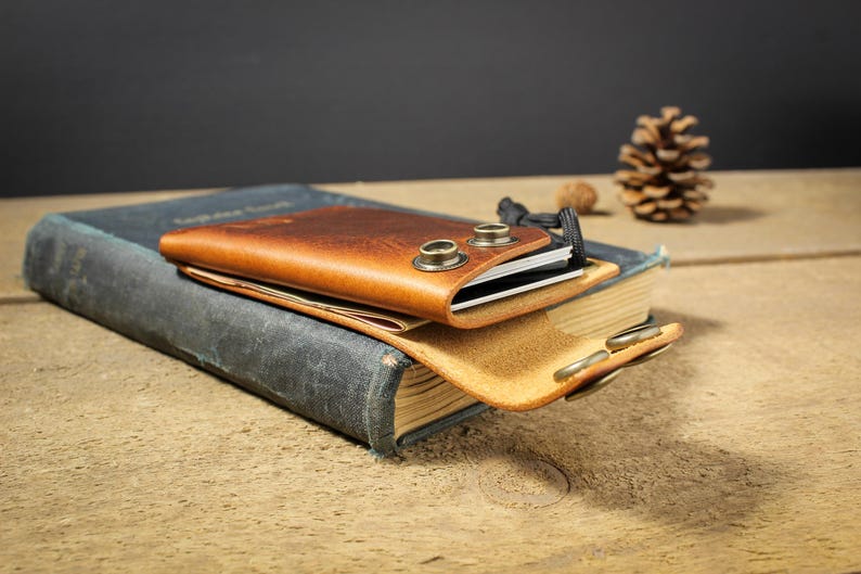 May include: A brown leather wallet with a snap closure and metal accents sits on top of a blue book. The wallet is open and has a black card holder inside.